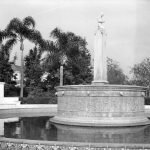 A black and white photograph of a circular fountain with a statue in the center surrounded by palm trees.
