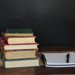 Stack of old books next to a spiral notebook and pen on a wooden table